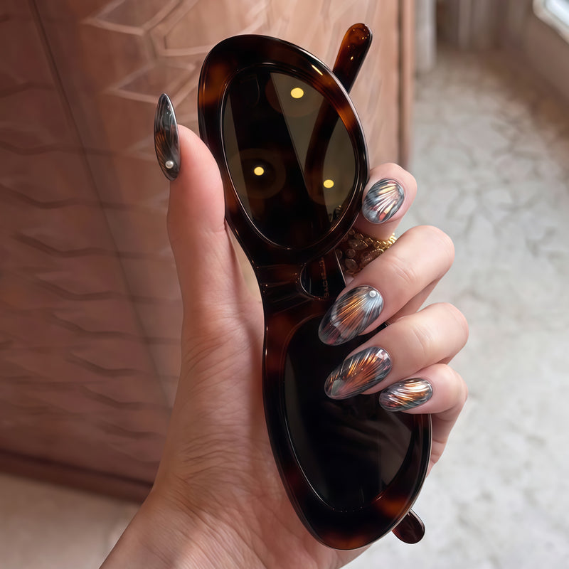 Hand holding oversized sunglasses while wearing black, gray, and copper handmade press-on nails with metallic fan patterns.