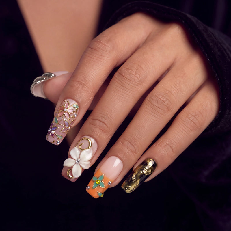 Hand model wearing ornate press-on nails with 3D florals, pearl accents, and gold charms on a dark background.