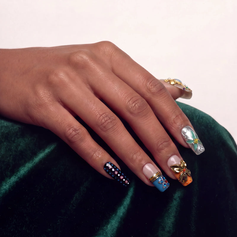 Hand model wearing ornate press-on nails with floral and gold accents on a dark green velvet background.