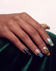 Hand model wearing ornate press-on nails with floral and gold accents on a dark green velvet background.