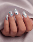 Close-up of a hand wearing silver-white press-on nails with blue rhinestones on a soft satin background.