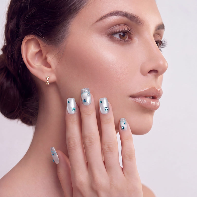 Hand model posing with silver-white press-on nails, blue rhinestones, and glitter tips against a white background.