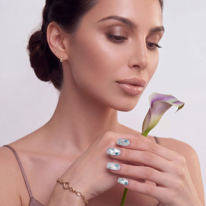 Hand model holding a lily stem while wearing silver-white press-on nails with blue rhinestones and glitter tips.