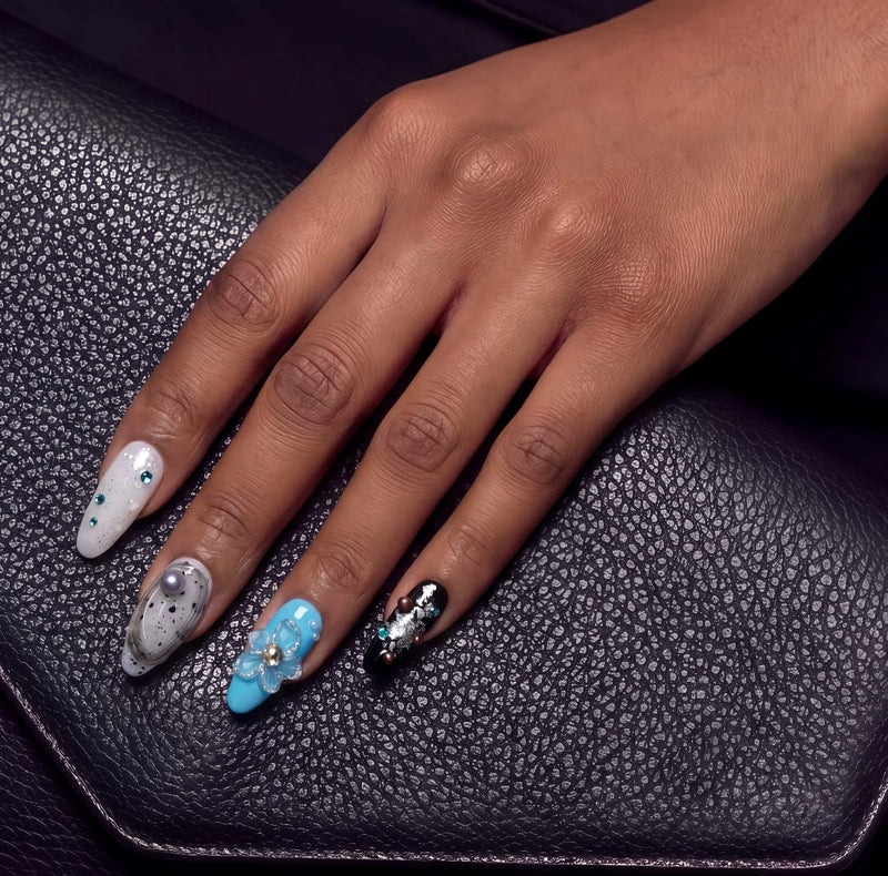 Hand wearing Artisian Orbit press-on nails with white, silver, blue, and black accents resting on a black leather surface