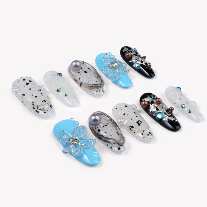 Diagonal flat lay of Artisian Orbit press-on nails with white, black, and blue accents, pearl details, and gem embellishments