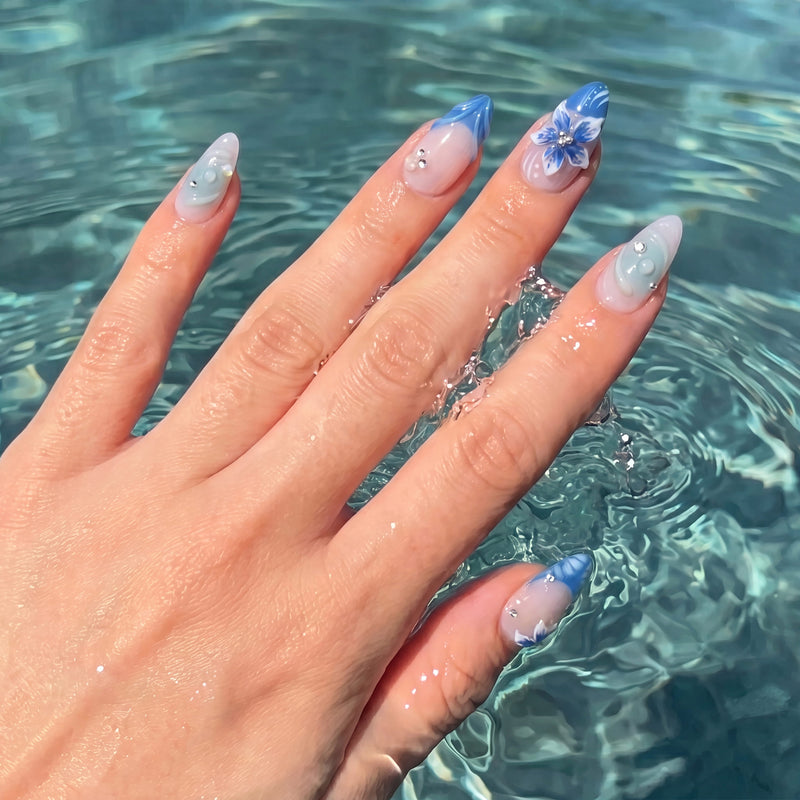 Hand model in pool water wearing Azure Bloom press-on nails with blue floral tips, aqua swirls, and silver rhinestones
