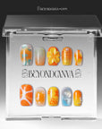 Clear acrylic packaging display showing the orange, yellow, blue, and gold press-on nail set inside a branded case.