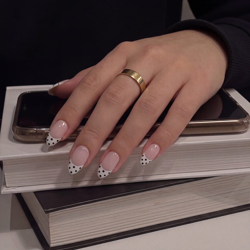Hand model wearing Bare Polka press-on nails in nude pink with black polka dot tips while resting on books and a phone