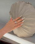 Hand model wearing Baroque Crown press-on nails with silver chrome, pearls, rhinestones, and white baroque accents in front of shell decor