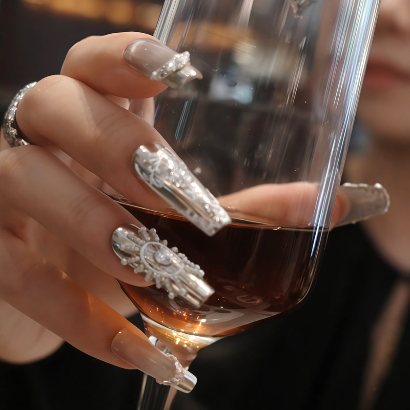 Hand model holding a wine glass while wearing Baroque Crown press-on nails with silver chrome, white baroque details, pearls, and rhinestones