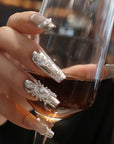 Hand model holding a wine glass while wearing Baroque Crown press-on nails with silver chrome, white baroque details, pearls, and rhinestones