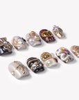Angled flat lay of Baroque Treasures press-on nails with black and ivory bases, burgundy heart detail, gold charms, pearls, and colorful gemstones