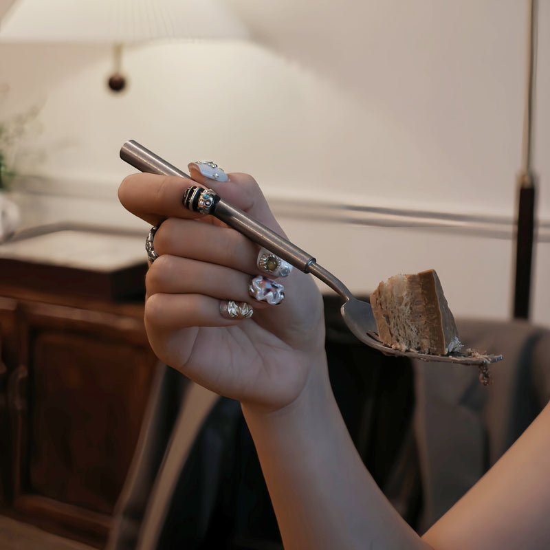 Lifestyle hand model holding a dessert fork while wearing Baroque Treasures press-on nails with pearl clusters, gold charms, and gemstone accents