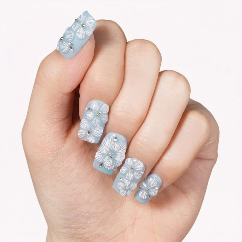 A hand model wearing light blue press-on nails with white floral petals and rhinestone accents