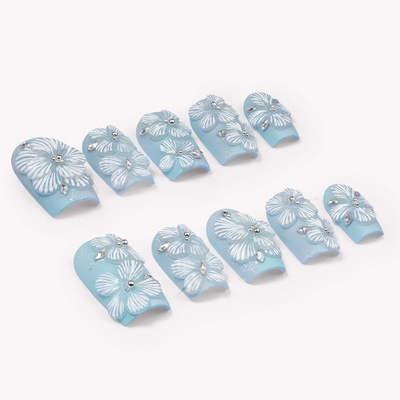 A set of light blue press-on nails with white floral petals and rhinestone accents on a white background