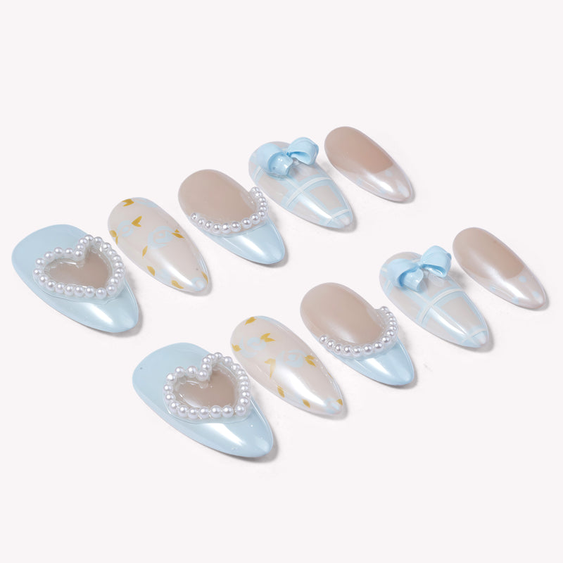 Diagonal flat lay of blue and nude press-on nails with pearl hearts, bows, pearl trim, and gingham accents on a white background.