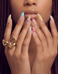 Hand model wearing multicolor floral press-on nails with pastel blue, pink, yellow, and burgundy details.