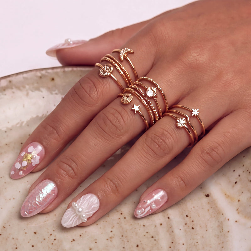 Hand close-up wearing blush pink press-on nails with gold rings stacked across the fingers.