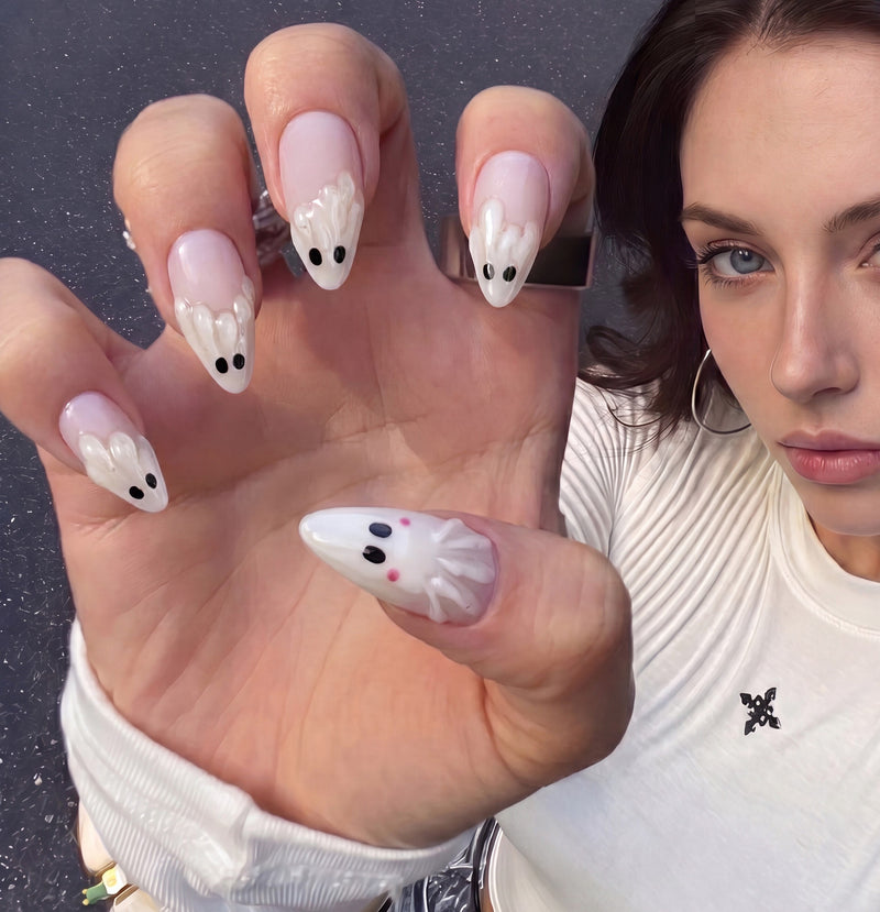 Close-up hand model wearing Boo Cuties press-on nails with pale pink bases, raised white ghost shapes, black eyes, and tiny pink cheeks