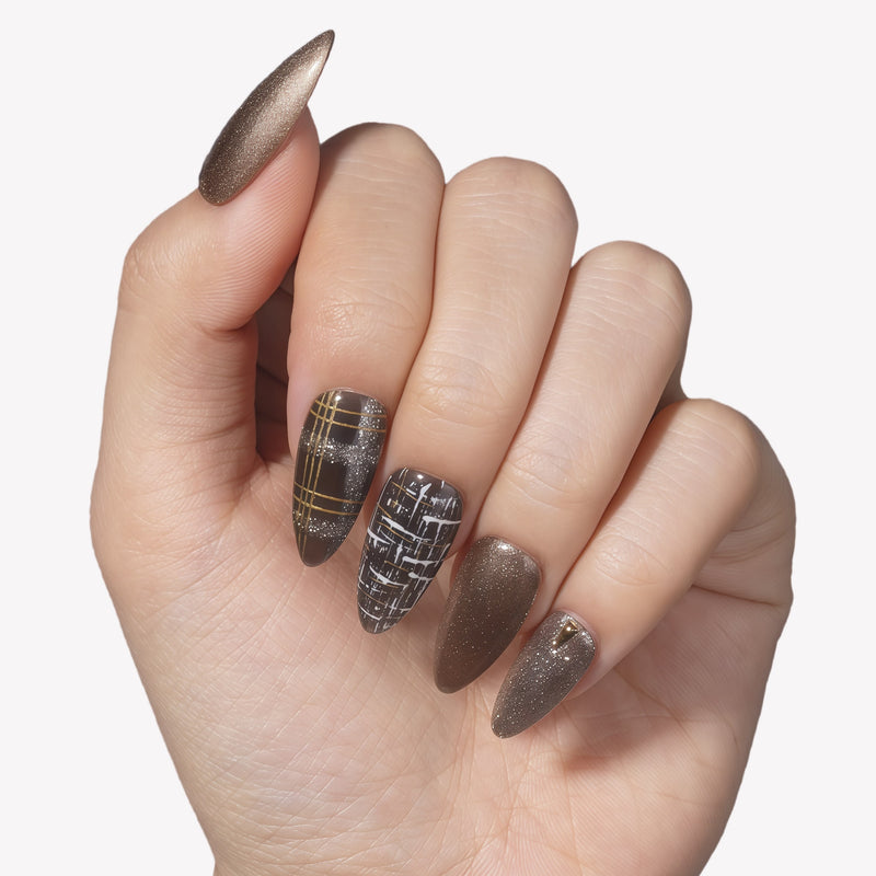 Hand model wearing bronzed press-on nails with black plaid, gold stripe, and glitter accents