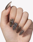 Hand model wearing bronzed press-on nails with black plaid, gold stripe, and glitter accents