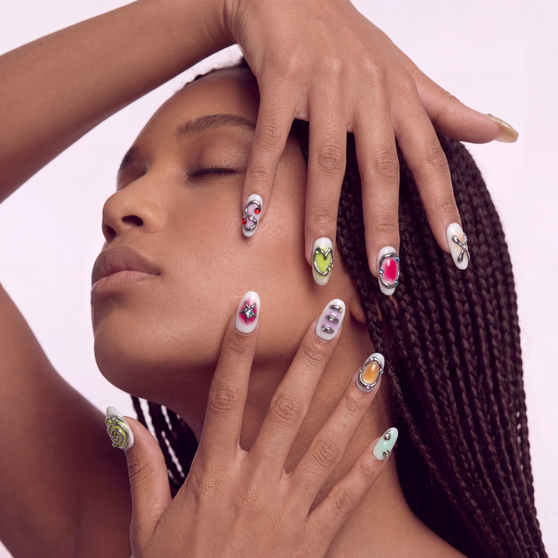 Face close-up hand model wearing Candy Chrome press-on nails with silver chrome outlines, pastel heart star and bow designs, glossy accents, and rhinestones