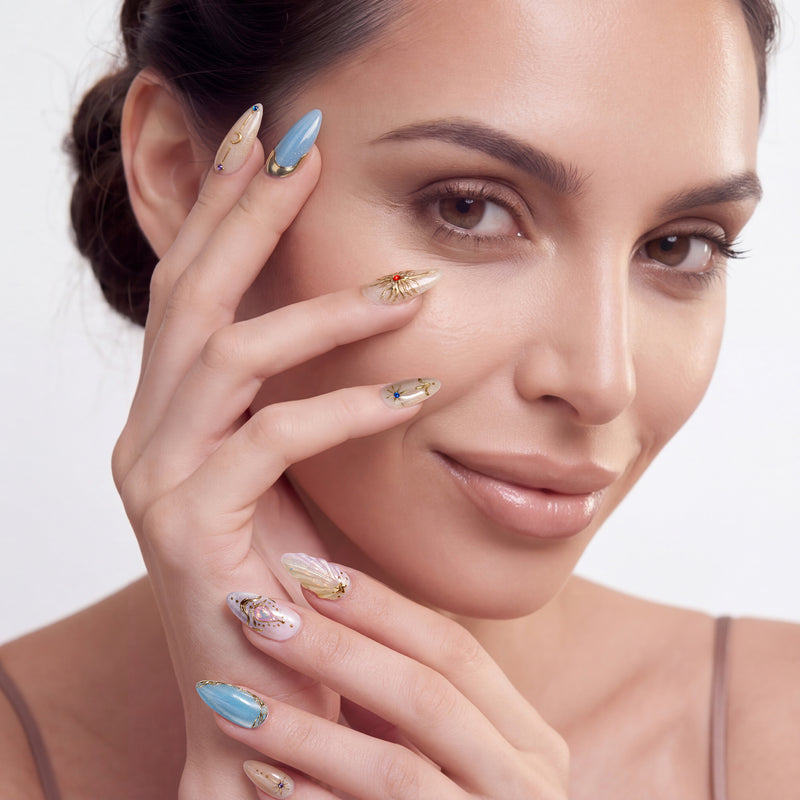Portrait of a hand model wearing blue and champagne almond press-on nails with gold embellishments and gem accents