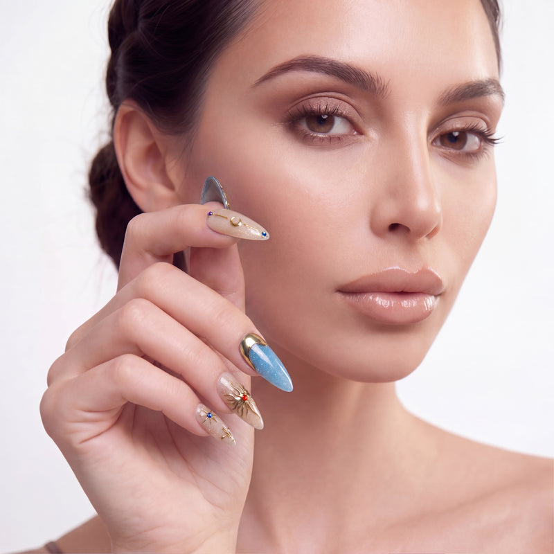 Portrait of a hand model wearing blue and champagne almond press-on nails with gold embellishments and gem accents