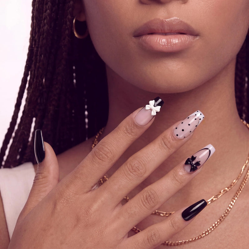 Face close-up hand model wearing Chérie Noir press-on nails with glossy black nails, nude pink bases, black polka dots, black French outline, and raised bow accents