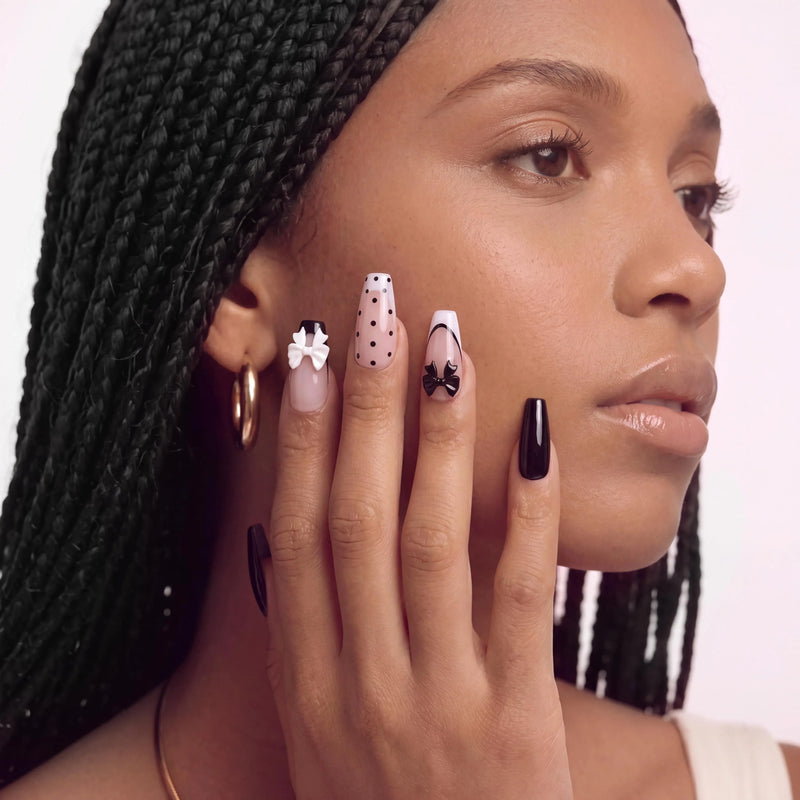 Side face close-up hand model wearing Chérie Noir press-on nails with glossy black nails, nude pink polka dots, black French outline, and raised black and white bow accents