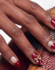 Close-up of a hand model wearing Cherry Devotion press-on nails in red, blush, and gold with cherries, bows, and hearts