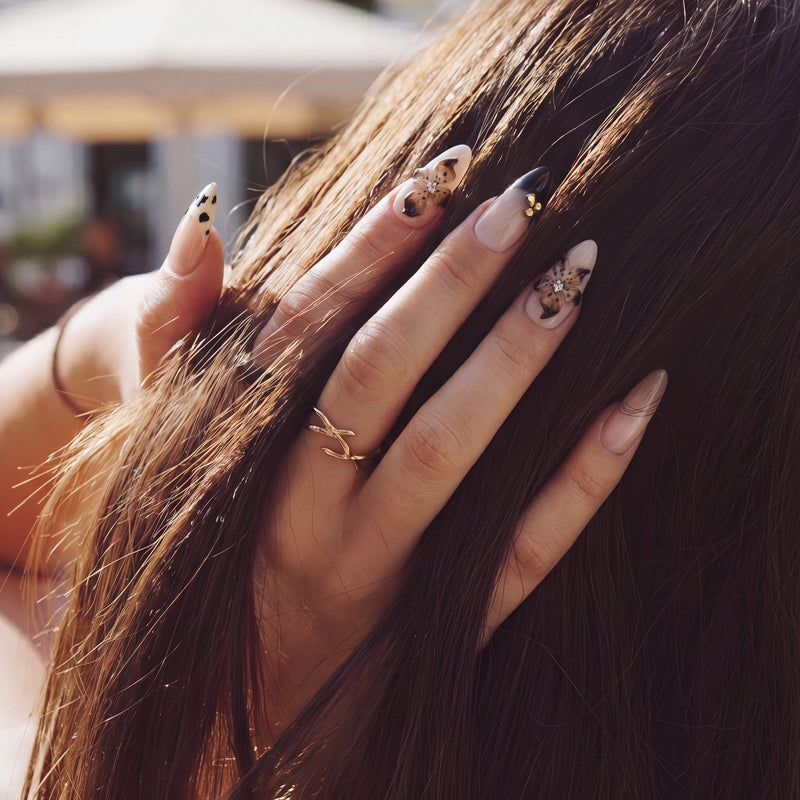 Hand model touching hair while wearing Chestnut Bloom press-on nails with nude pink bases, brown floral petals, black French tips, and gold accents