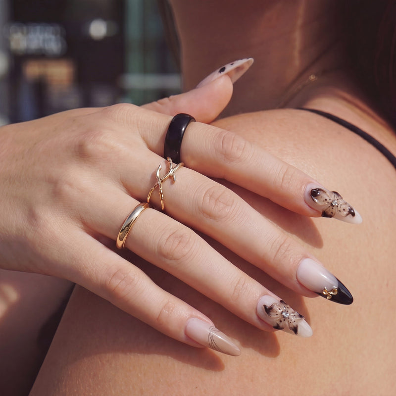 Lifestyle hand model wearing Chestnut Bloom press-on nails with nude pink bases, brown floral petals, black French tips, and gold ring styling