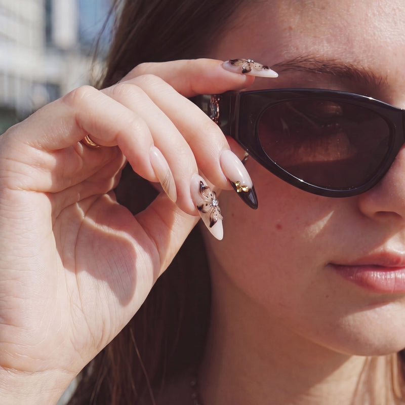 Close-up hand model wearing Chestnut Bloom press-on nails with brown floral petals, black French tips, and rhinestone accents beside sunglasses