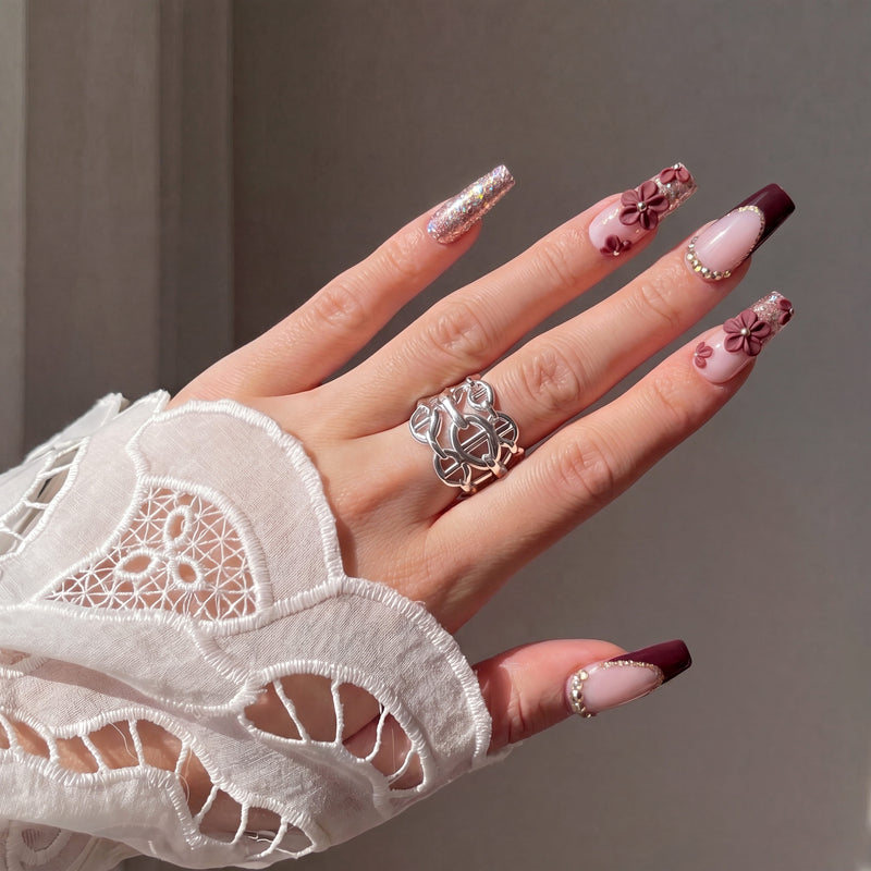 Close-up hand model wearing Coffee Blossom press-on nails with brown French tips, glitter, rhinestones, and 3D flowers beside a lace sleeve