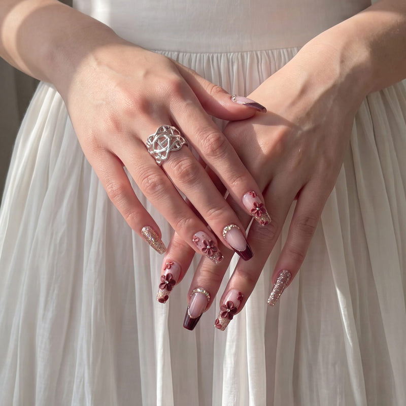 Hand model wearing Coffee Blossom press-on nails with brown French tips, 3D floral accents, rhinestones, and glitter over a white dress