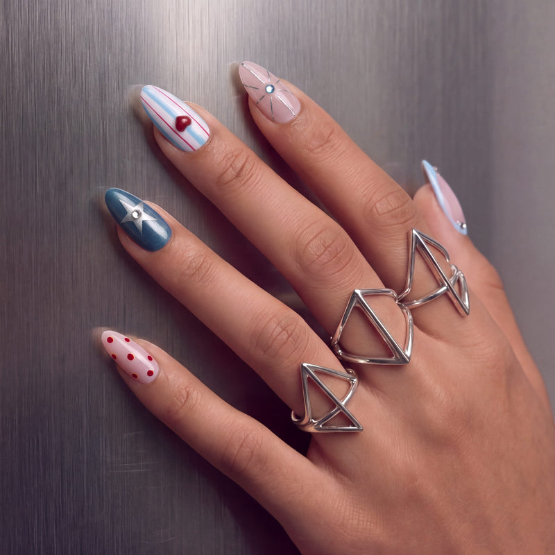 Hand model wearing Cosmic Candy press-on nails with blue and white stripes, red heart charm, silver star accent, red polka dots, rhinestones, and silver rings