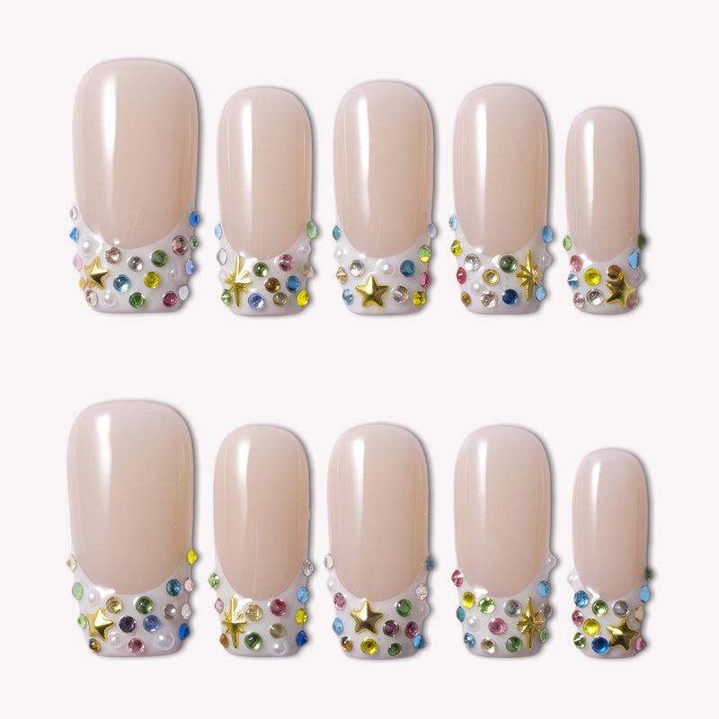 Flat lay of nude French tip press-on nails with multicolor rhinestones, pearls, and gold star charms on a white background