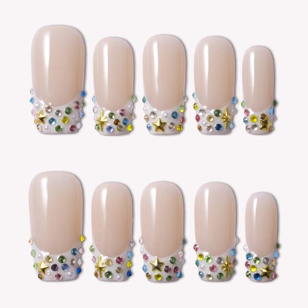 Flat lay of nude French tip press-on nails with multicolor rhinestones, pearls, and gold star charms on a white background