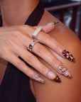 Hand model wearing pale pink and burgundy press-on nails with gold swirls, crystals, and cross embellishments.