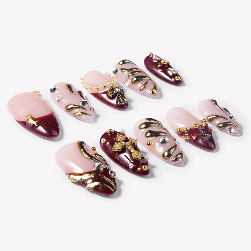 Flat lay of burgundy and pale pink press-on nails with gold swirls, rhinestones, pearls, and cross charms