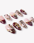 Flat lay of burgundy and pale pink press-on nails with gold swirls, rhinestones, pearls, and cross charms
