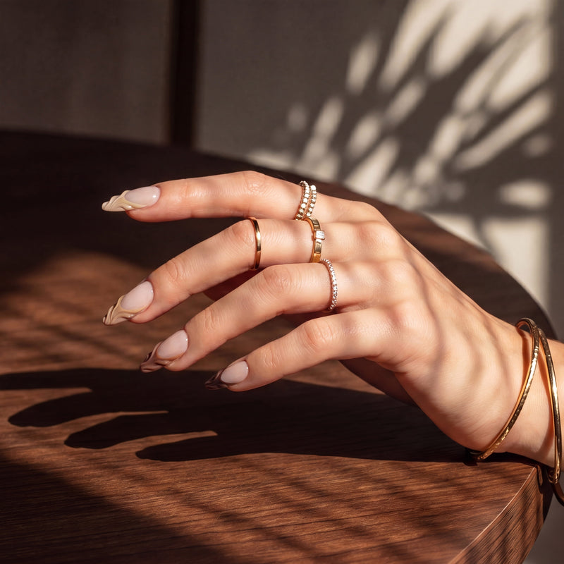 Hand model wearing Dance of Shifting Sands press-on nails with nude bases and sculpted sand wave tips styled with gold jewelry on a wood table