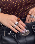 Hand model holding a black bag while wearing Dark Masquerade press-on nails with glossy black tips, pink rhinestone accents, and fishnet details