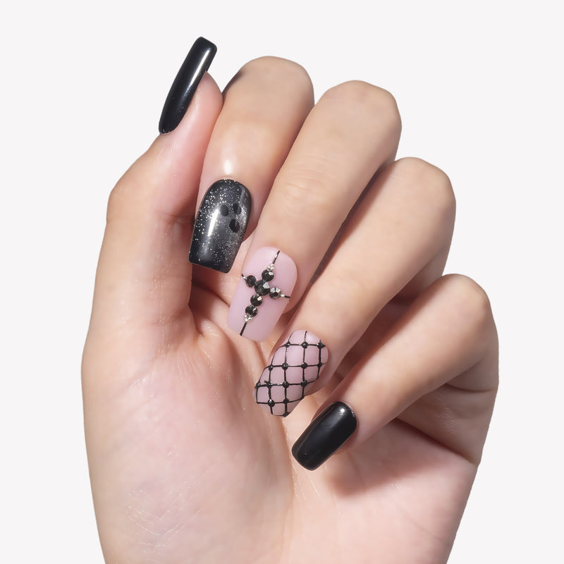 White background hand model wearing Dark Masquerade press-on nails with glossy black nails, black glitter accents, pink rhinestone cross, and fishnet pattern