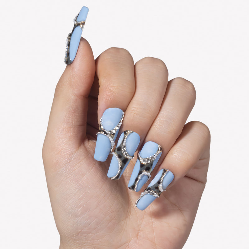 White background hand model wearing Denim Luxe press-on nails with sky blue bases, gold black leopard accents, pearl chains, and rhinestones