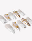 Angled flat lay of Divine Grace press-on nails with white sculpted bases, nude pearl shimmer, gold chrome accents, bead details, and rhinestone crosses