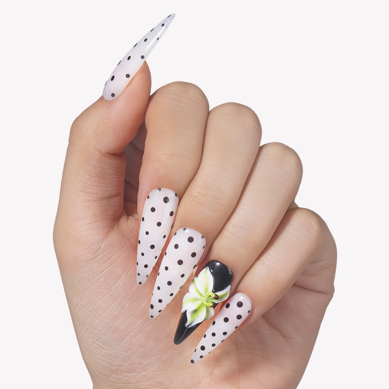 Hand model wearing Eden Polka press-on nails with white bases, black polka dots, glossy black accent nail, and raised green white flower design