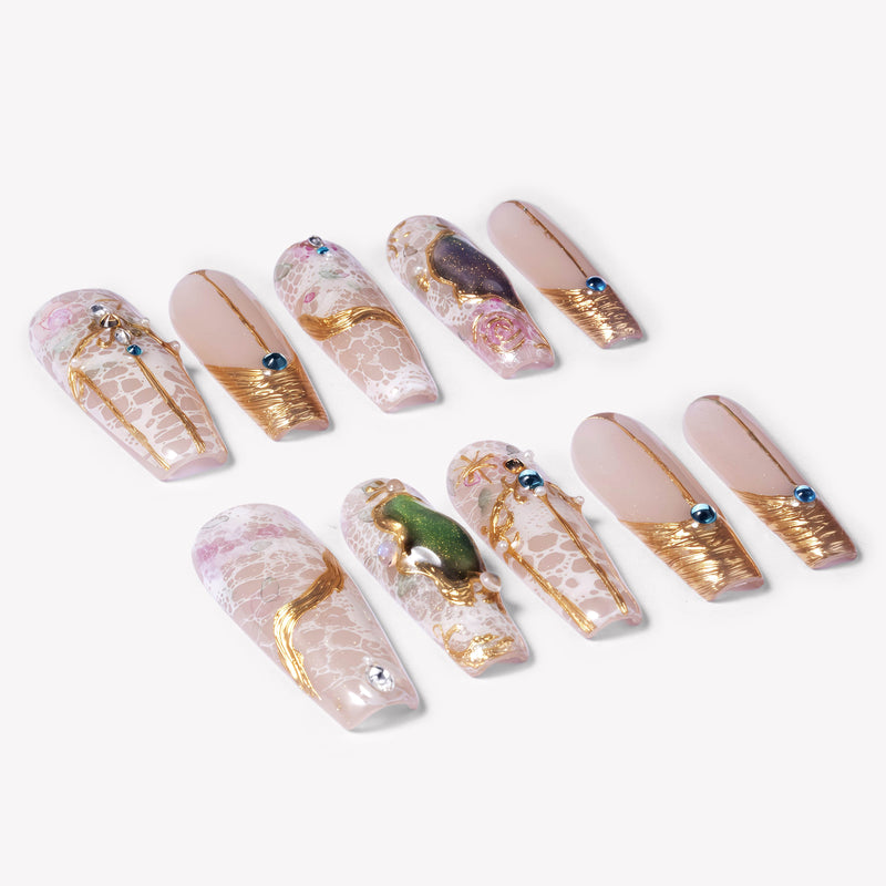 Angled flat lay of Enchanted Garden press-on nails with pale pink translucent bases, raised gold linework, blue rhinestones, and green gemstone charms
