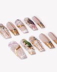 Angled flat lay of Enchanted Garden press-on nails with pale pink translucent bases, raised gold linework, blue rhinestones, and green gemstone charms